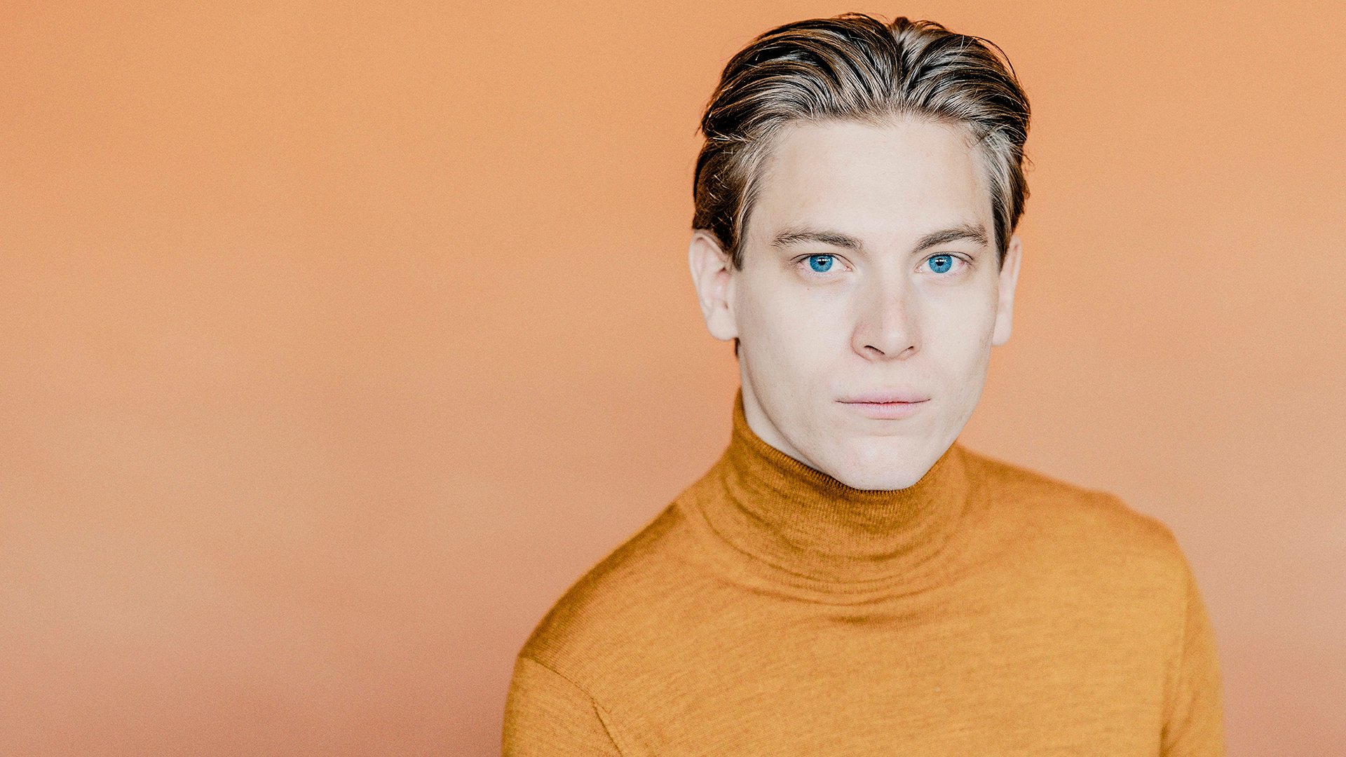 Klaus Makela mit kurzem Haar und Rollkragenpullover, vor einfarbigem orangefarbenem Hintergrund.
