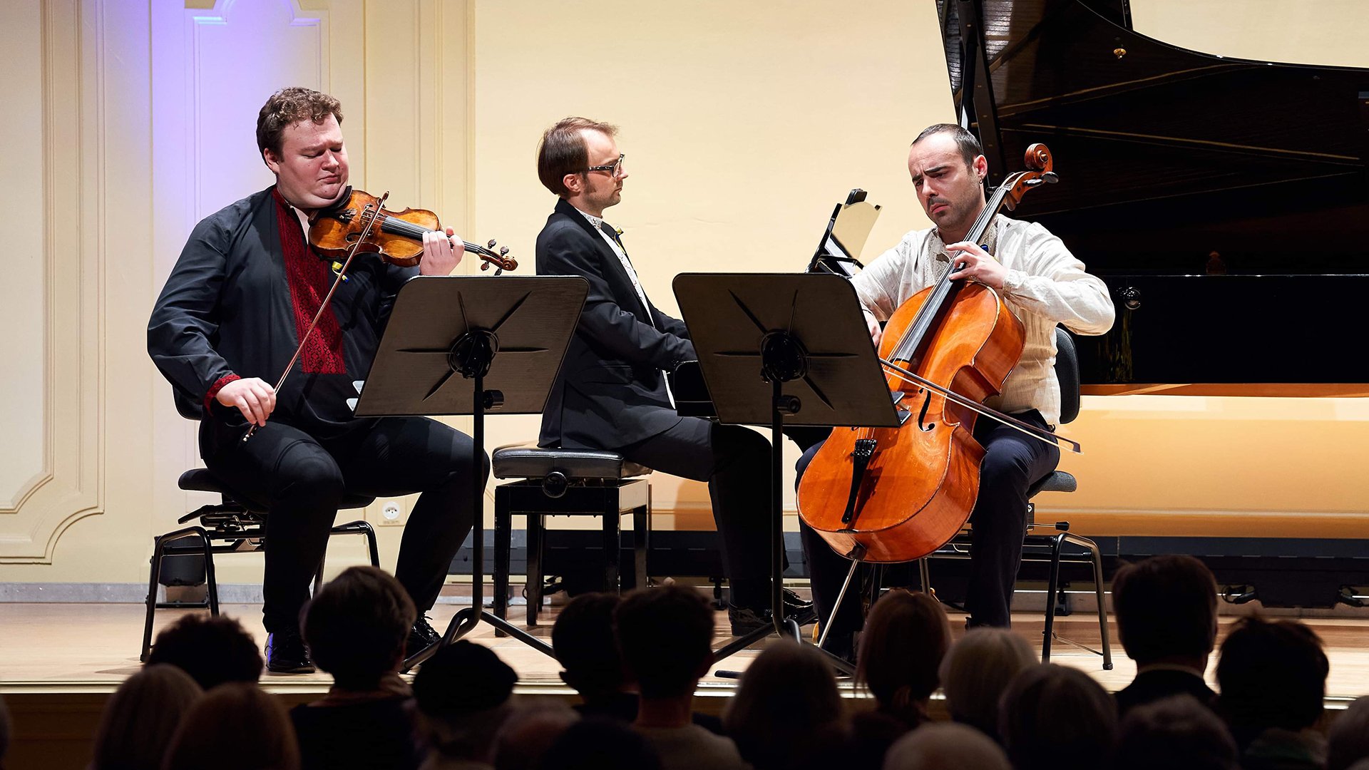 Drei Musiker bei einem Kammerkonzert auf einer Bühne: Violine, Klavier und Violoncello. Im Vordergrund ist das Publikum zu sehen, im Hintergrund ein Konzertflügel.