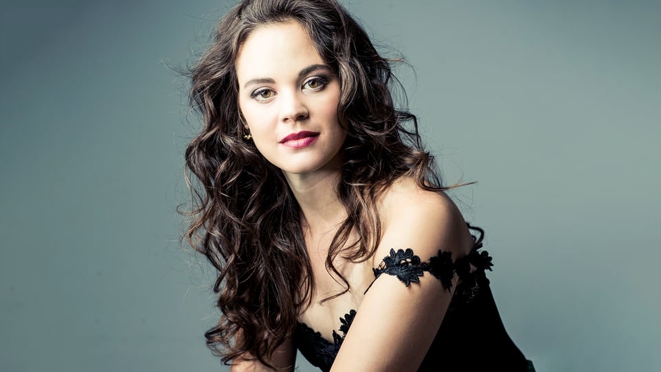 Porträt der Sopranistin Regula Mühlemann mit lockigem Haar und schwarzem Kleid vor hellem Hintergrund.