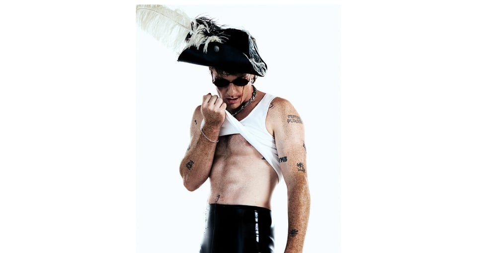 Mann mit schwarzem Piratenhut, weißer Feder und Sonnerbrille, das Unterhemd hochziehend 