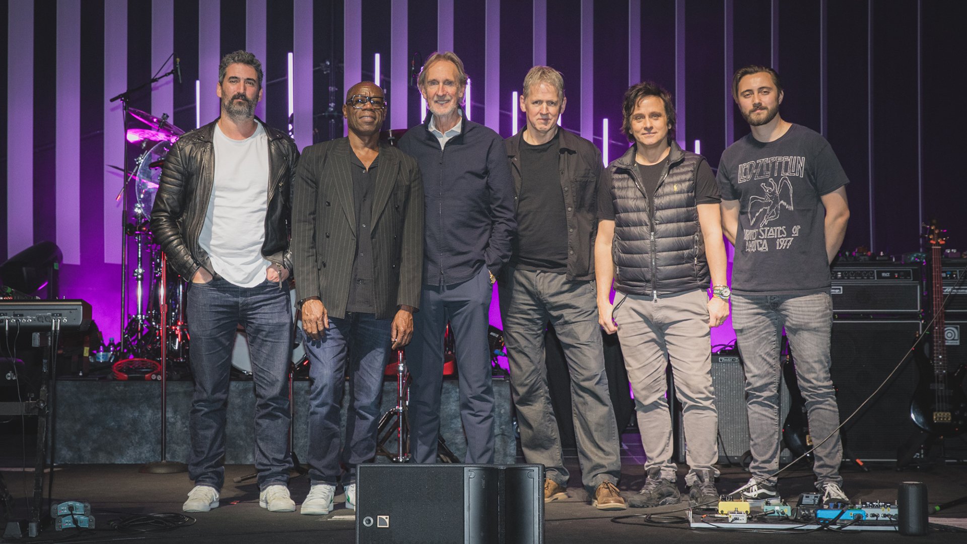 Bandfoto von Mike and The Mechanics mit sechs Bandmitgliedern auf einer Konzertbühne vor Schlagzeug, Verstärkern und violett beleuchteten Lichtstäben im Hintergrund