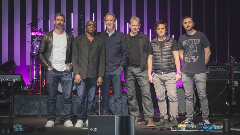 Bandfoto von Mike and The Mechanics mit sechs Bandmitgliedern auf einer Konzertbühne vor Schlagzeug, Verstärkern und violett beleuchteten Lichtstäben im Hintergrund