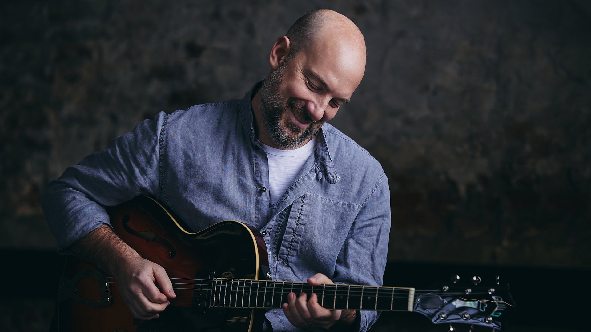 Person spielt eine E‑Gitarre vor neutralem Hintergrund, nach unten blickend, in heller Kleidung und mit Nahaufnahme der Hände auf den Saiten.