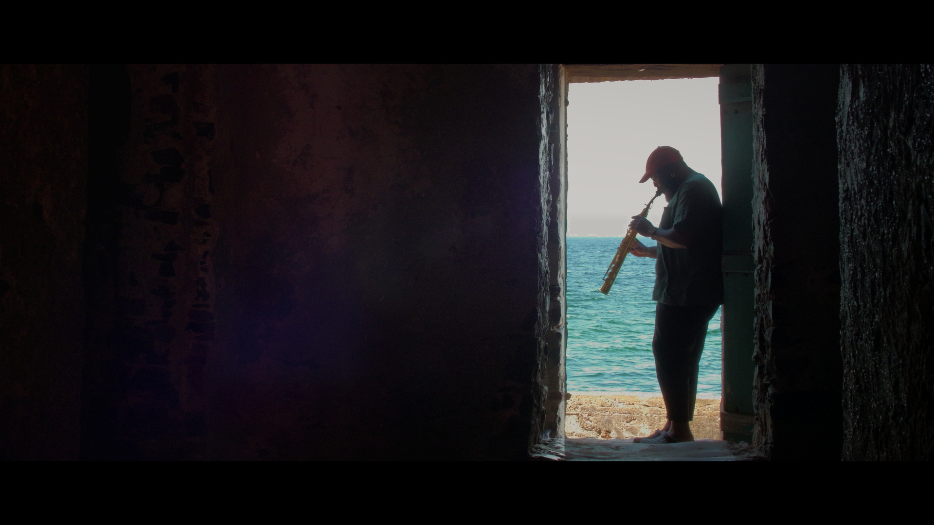 Filmszene aus Tukki mit einer silhouettenhaft dargestellten Person, die in einem Türdurchgang am Meer ein Blasinstrument spielt, vor hellem Tageslicht und Wasser im Hintergrund, eingerahmt von einem dunklen Innenraum.