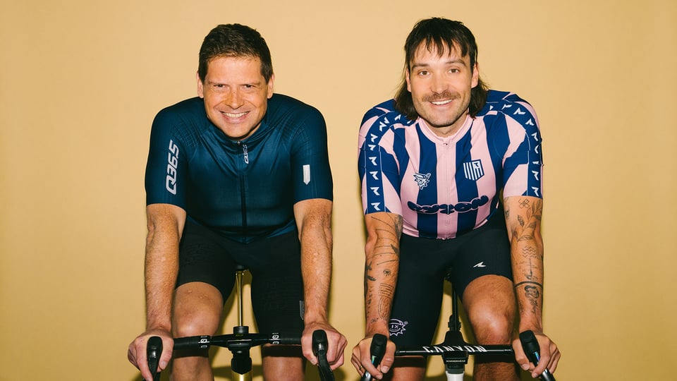 Jan Ullrich und Rick Zabel lachend nebeneinander auf Fahrrädern in Sportkleidung, vor gelbem Hintergrund