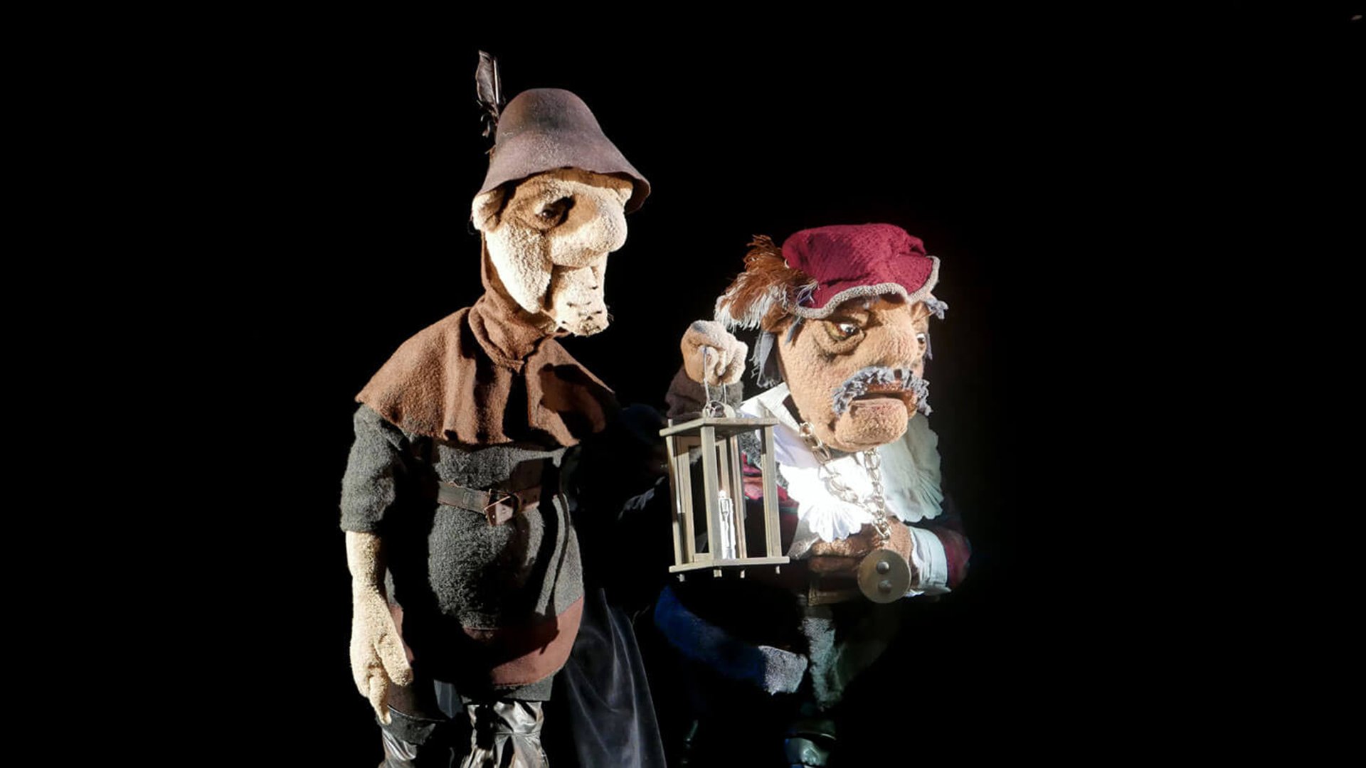 Zwei detailreich gestaltete Figurenpuppen auf dunkler Bühne; eine hält eine Laterne, beide tragen historische Kostüme und sind von oben beleuchtet vor schwarzem Hintergrund.