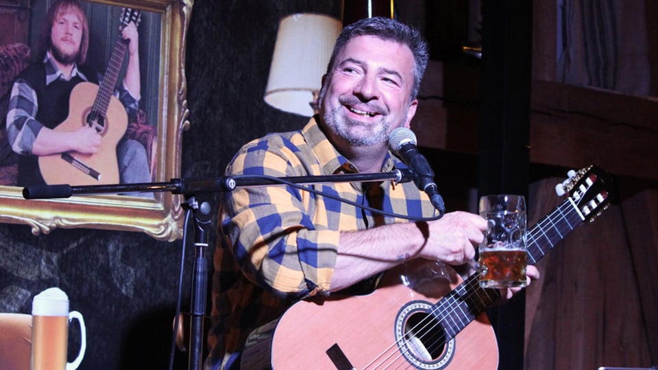 Bühnenfoto eines Musikkabarett‑Auftritts: Eine Person sitzt auf einer kleinen Bühne mit einer Akustikgitarre und spricht in ein Mikrofon. In der rechten Hand wird ein Bierkrug gehalten, die Gitarre liegt auf dem Schoß. Im Hintergrund ist ein gerahmtes Porträt einer weiteren guitarrespielenden Person an der Wand zu sehen sowie gedämpfte Bühnenbeleuchtung. Die Szene vermittelt eine intime Kleinkunst‑Atmosphäre mit Musik, Sprache und humorvollem Bühnenformat.