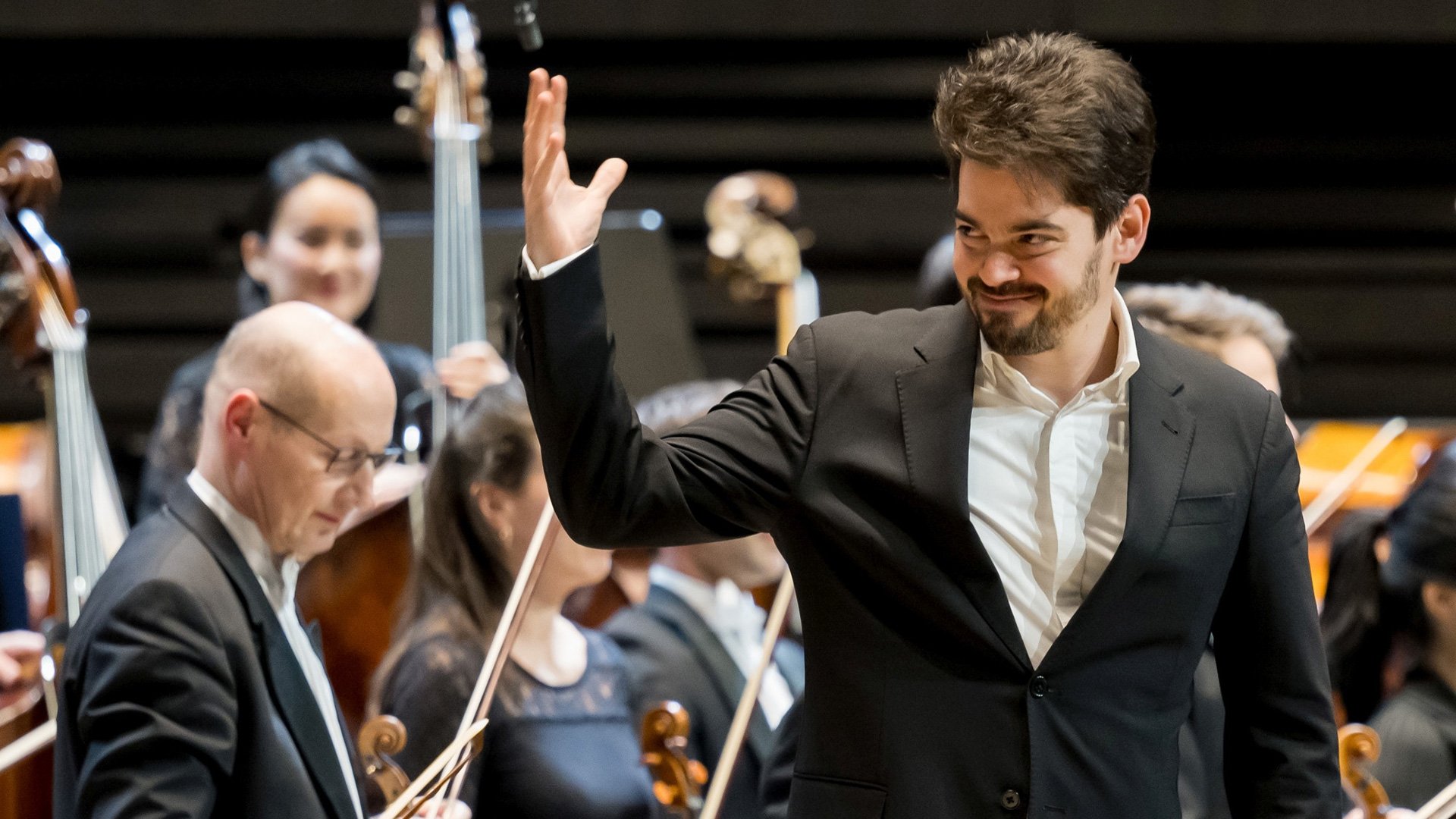 Foto von Lahav Shani, wie er bei einem Konzert grüßt, mit dem Orchester im Hintergrund.