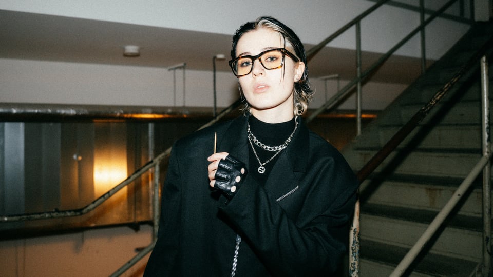 Foto für die Veranstaltung NESS – HAUTNAH TOUR (AKUSTIK) 2026 mit der Künstlerin, die eine Brille trägt, und einer Treppe im Hintergrund