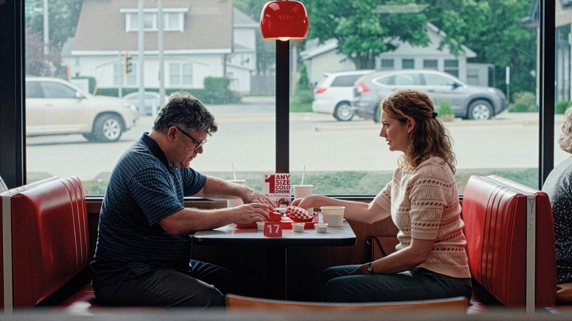 Zwei Personen sitzen sich in einem Diner gegenüber an einem Tisch mit Essen und Getränken, große Fenster im Hintergrund.
