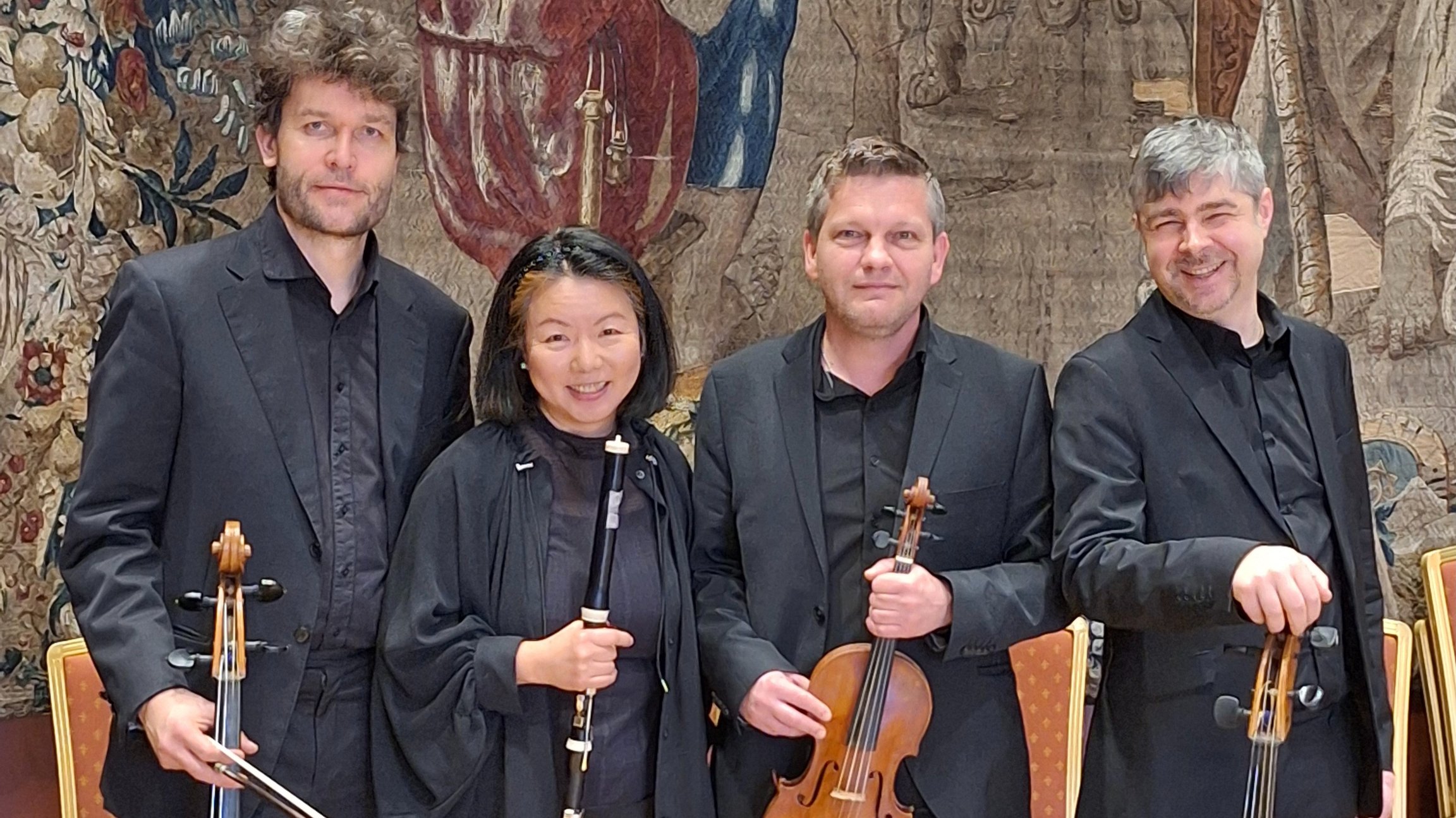 Vier Musikerinnen und Musiker in schwarzer Konzertkleidung stehen mit Celli, Klarinette und Violine vor einem historischen Wandteppich in einem Konzertsaal und blicken in die Kamera.