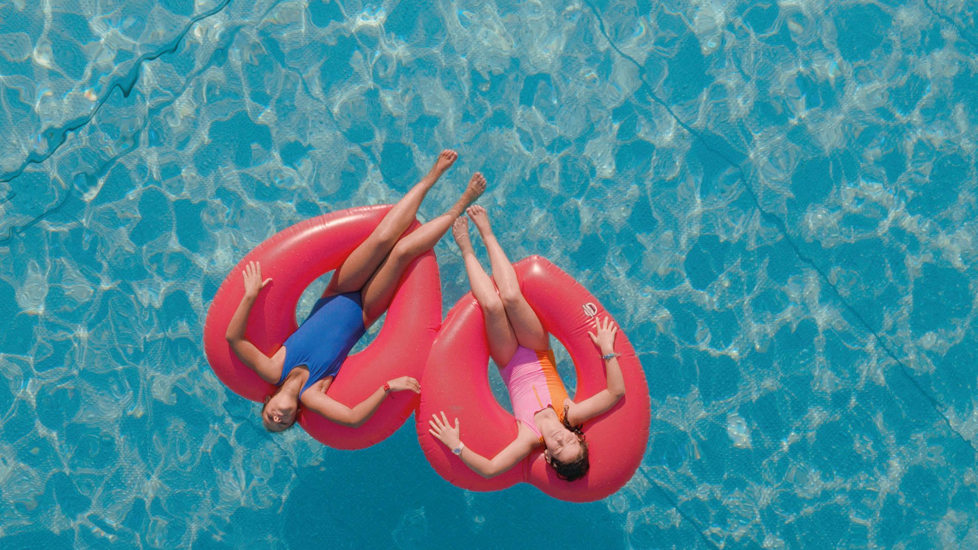Zwei Personen liegen nebeneinander auf einer großen, roten aufblasbaren Insel im Wasser eines Pools; das Wasser ist klar und blau, und die Szene ist aus der Vogelperspektive aufgenommen, sodass die Körper parallel ausgerichtet auf der Wasseroberfläche treiben.