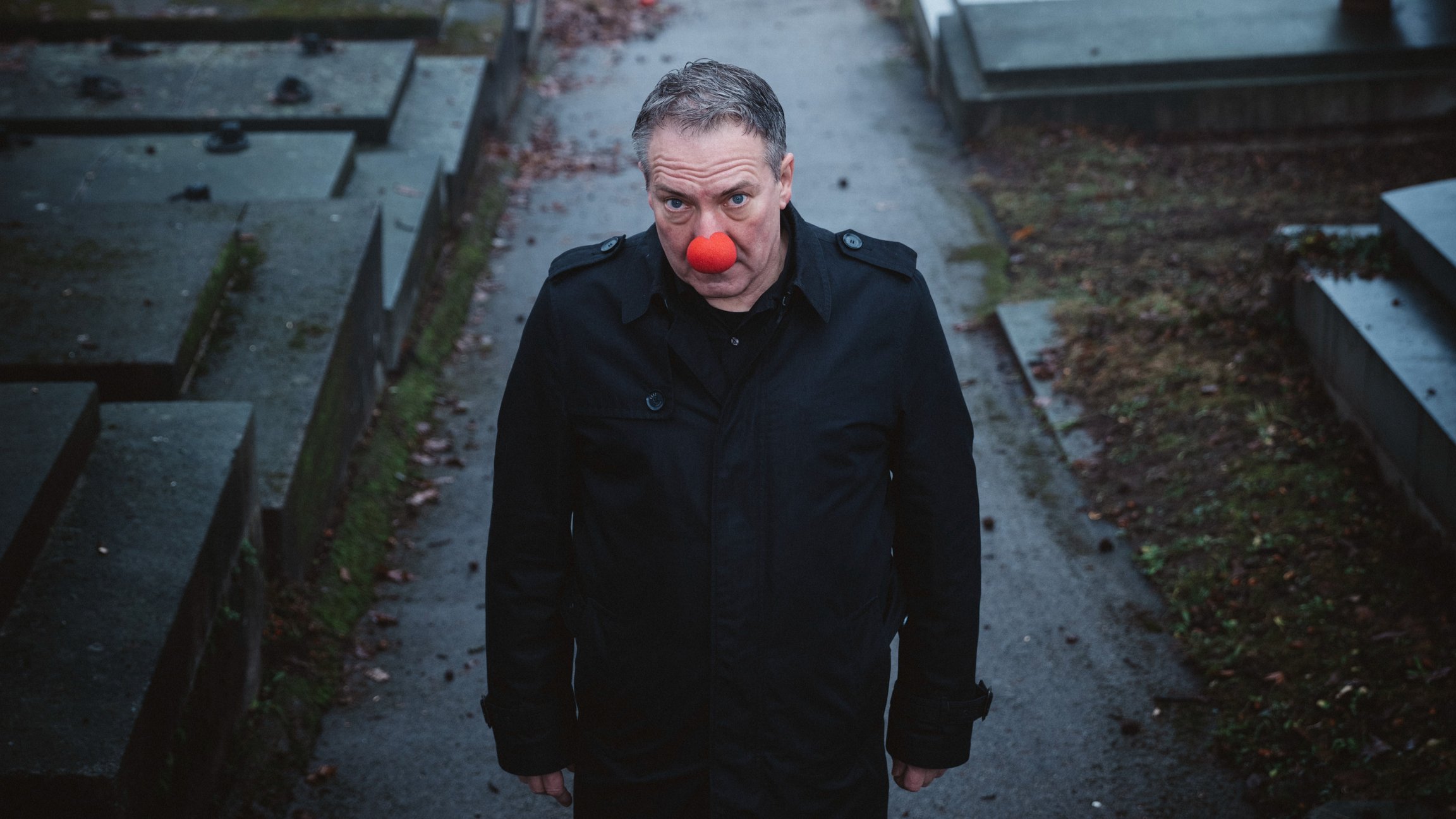 Robert Palfrader mit einer roten Clownsnase in einem schwarzen Mantel auf einem dunklen Weg, links und rechts schwarze  Steinplatten