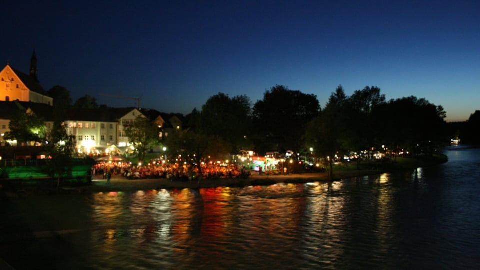 Abendliche Uferszene an einem Fluss: Entlang des beleuchteten Uferbereichs sitzen zahlreiche Besucherinnen und Besucher an Tischen bei einem Nachtflohmarkt oder Abendmarkt; Lichter von Ständen und Gebäuden spiegeln sich im Wasser. Im Hintergrund sind Bäume, historische Häuser und eine Kirche zu erkennen, darüber ein dunkler Abendhimmel. Das Bild vermittelt sommerliche Atmosphäre, Marktgeschehen und Stadtleben bei Nacht.