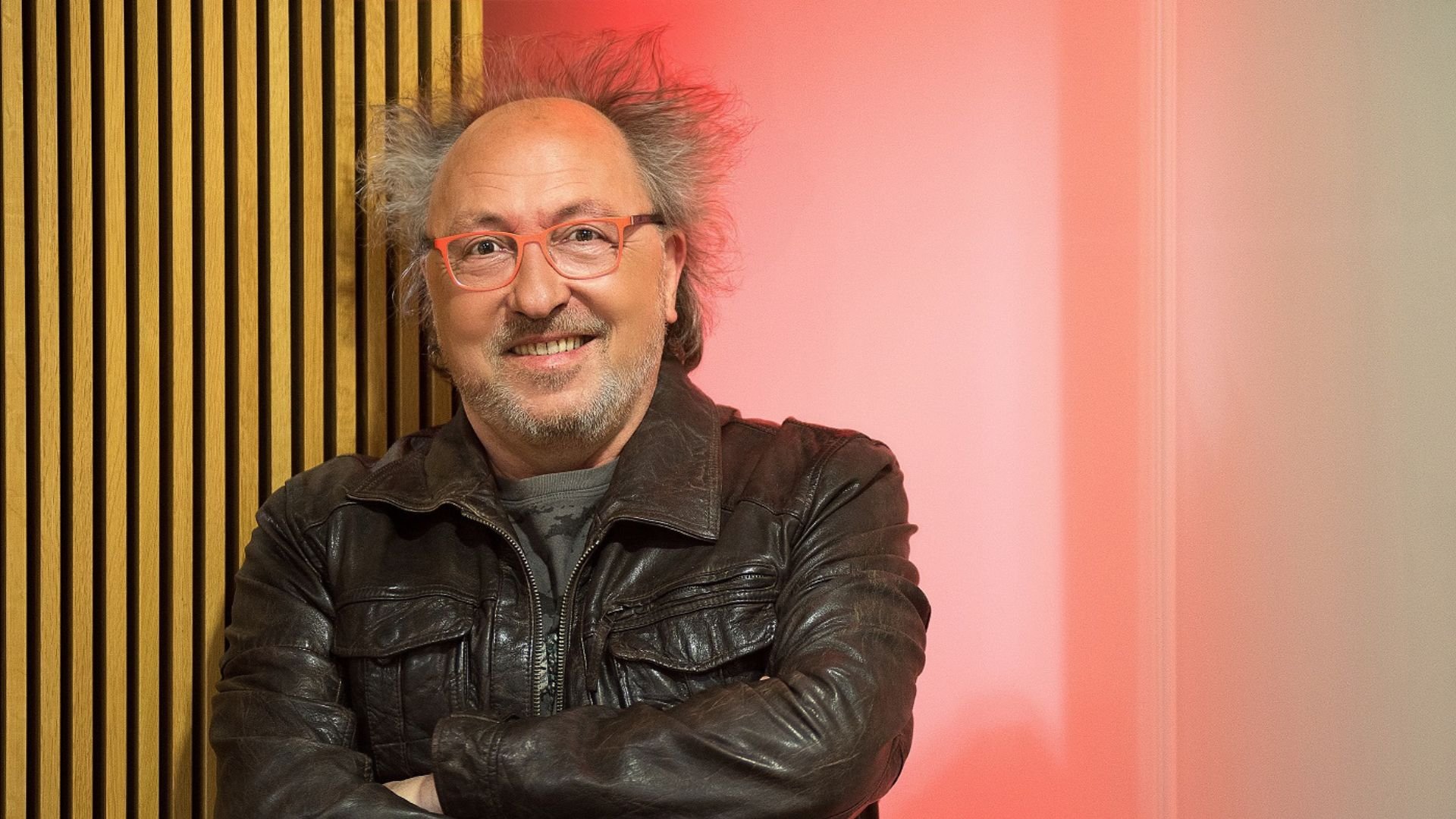 Dr Kabarettist Urban Priol in einer dunklen Lederjacke steht mit verschränkten Armen vor einer Wand mit Holzpaneelen und roter Beleuchtung