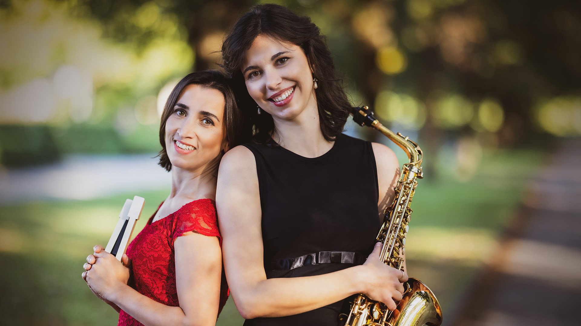 Zwei Musikerinnen stehen Rücken an Rücken in einer Parkanlage. Eine Person hält ein Saxofon, die andere Notenblätter; beide tragen elegante Kleidung vor unscharfem, grünem Hintergrund.