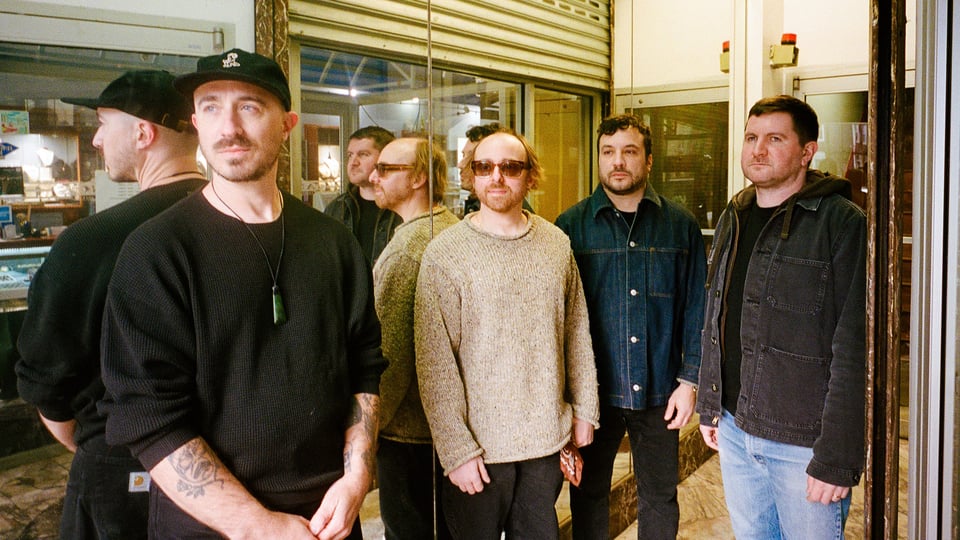 Bandfoto von The Menzingers mit vier Bandmitgliedern in Freizeitkleidung vor einer Schaufensterfront mit Spiegelungen, darunter eine Person mit schwarzer Kappe, eine mit Sonnenbrille und drei weitere in Pullovern und Jacken