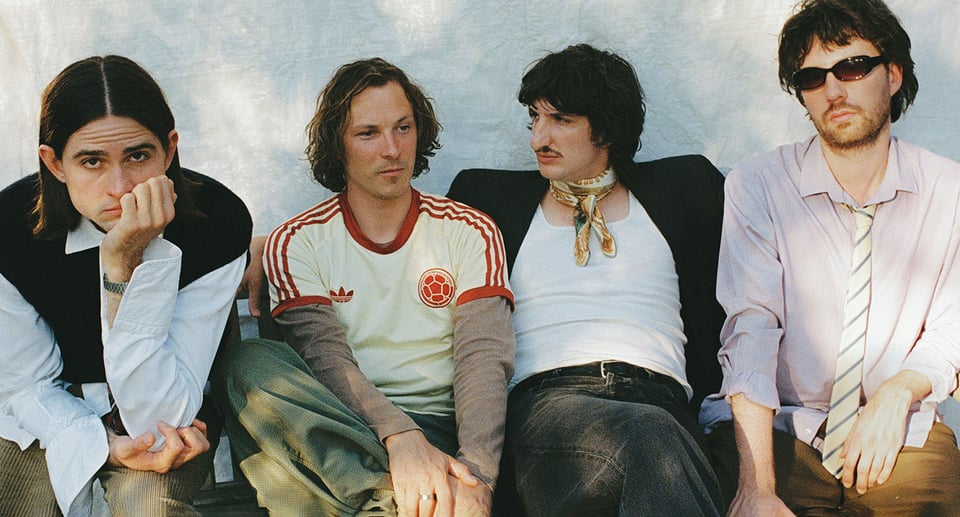Ein Foto der Band, sitzend auf einer Bank, mit einer weißen Wand im Hintergrund