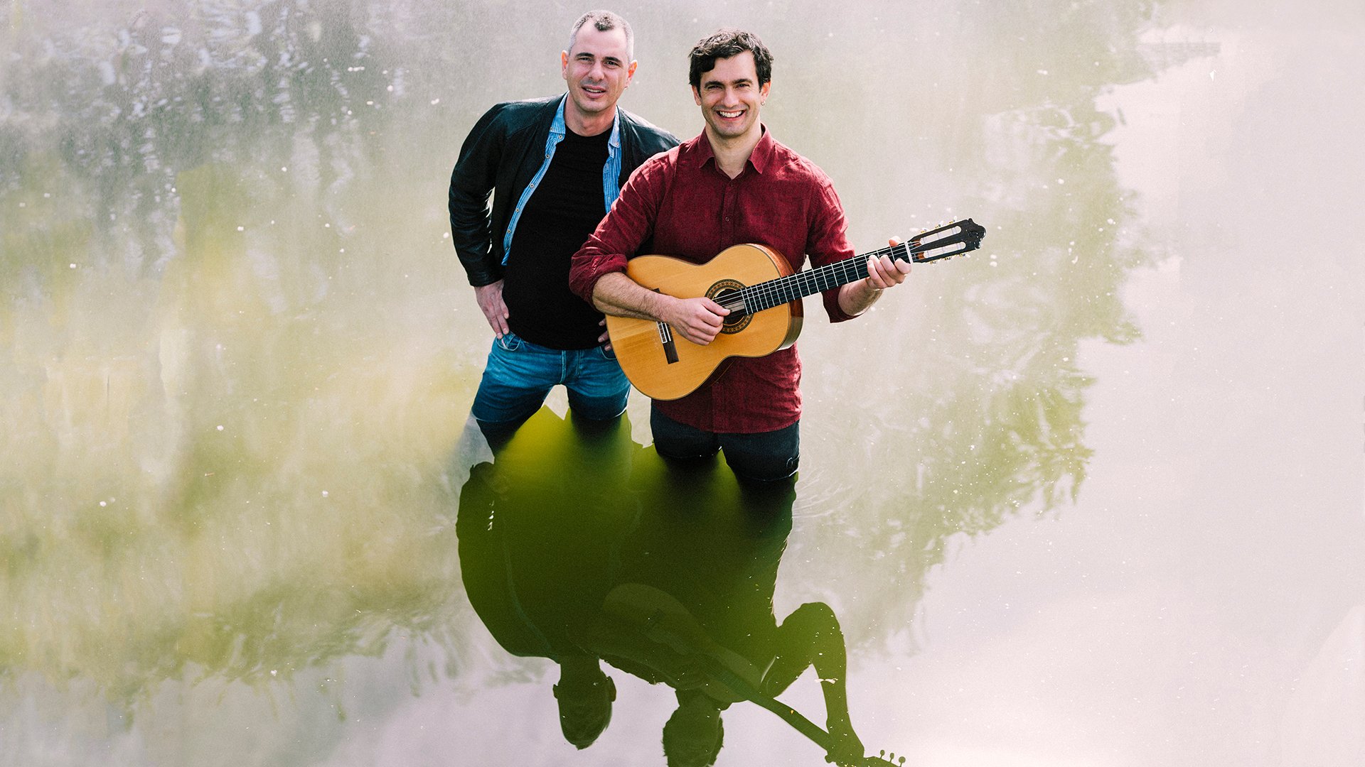 Zwei Personen stehen bis zur Hüfte im Wasser; eine hält eine akustische Gitarre, während ihre Spiegelbilder deutlich auf der Wasseroberfläche zu sehen sind