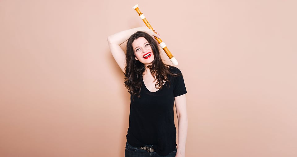 Dame mit dunklen langen Haaren und schwarzer kleidung mit Instrument vor rosa Hintergrund