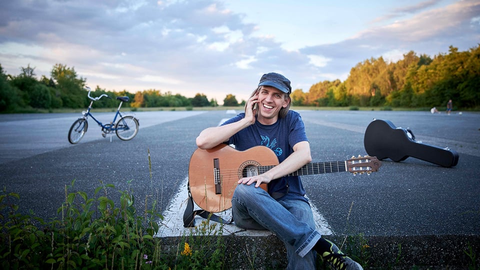 Porträtfoto von Weiherer mit Akustikgitarre auf einer breiten Asphaltfläche, sitzend vor Fahrrad und Gitarrenkoffer, umgeben von Wiesen und Bäumen im warmen Abendlicht