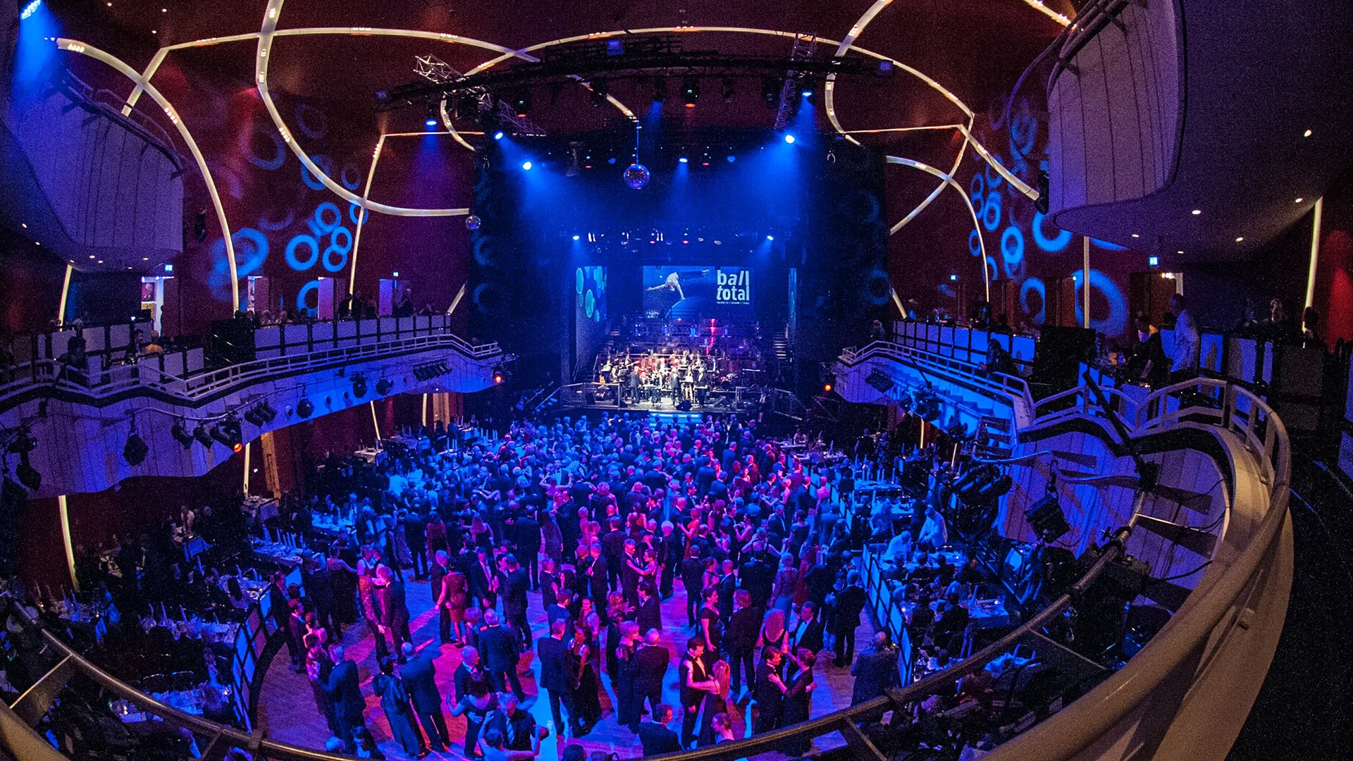 Ein Foto für das Event Ball Total 2026, mit dem Deutschen Theater von innen, tanzenden Menschen und einer Leinwand im Hintergrund mit blauen Lichtern.