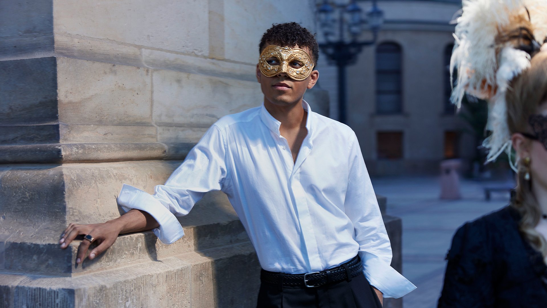 Louis Philippson Person in weißem Hemd mit goldfarbener Venezianischer Maske, an eine steinerne Gebäudefassade gelehnt, vor historischer Stadtkulisse, im Vordergrund sind Teile weiterer Maskenkostüme sichtbar