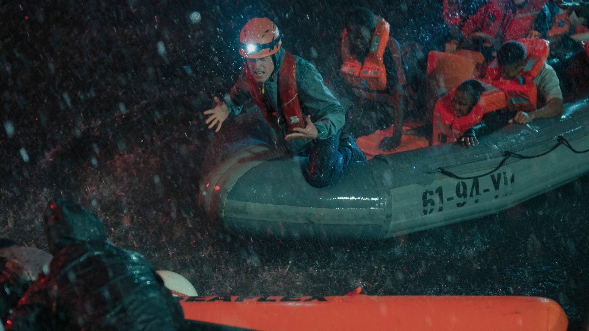 Eine Gruppe von Personen sitzt mit Rettungswesten in einem Rettungsboot im Wasser vor einem weiteren Rettungsboot während es stark regnet.