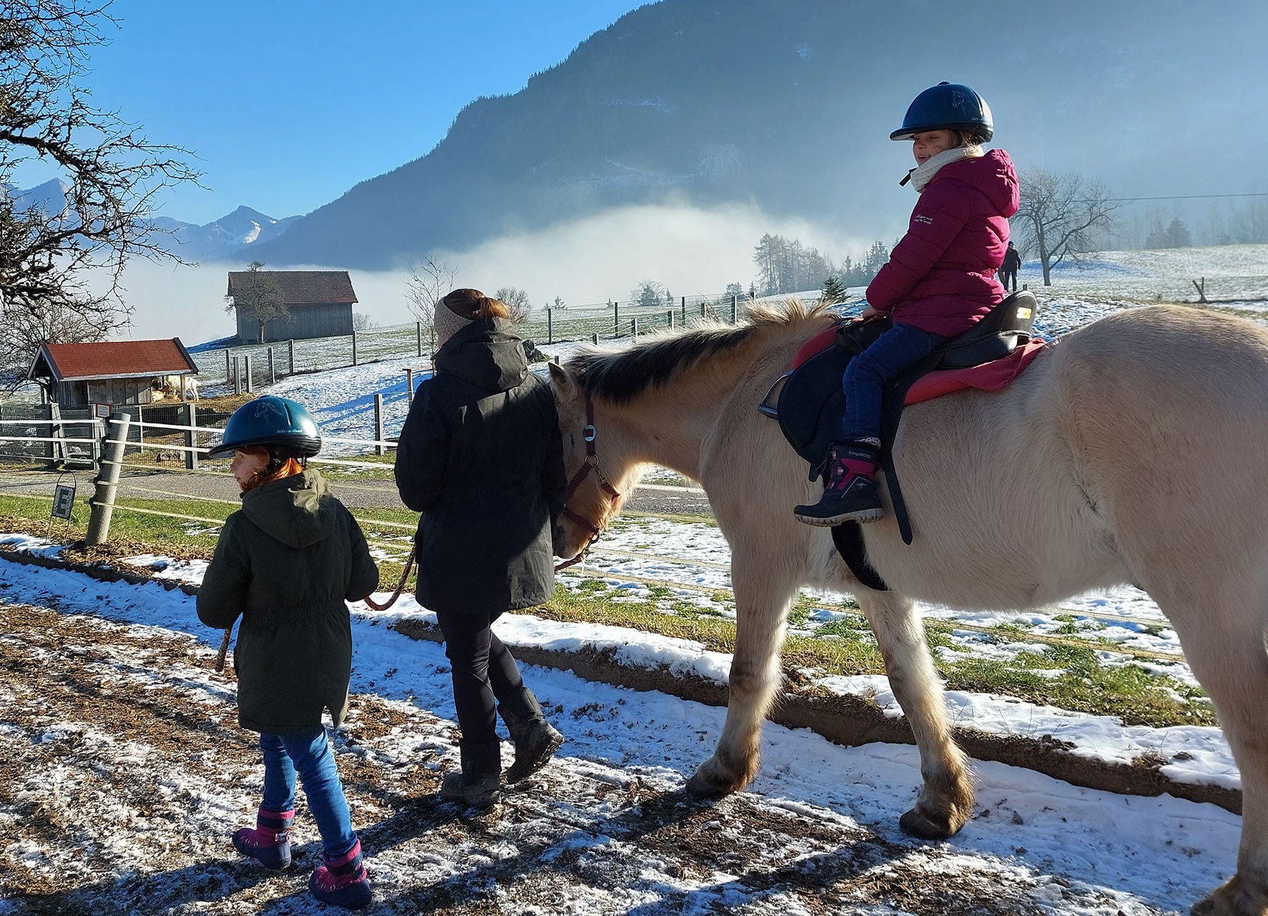 Foto von zwei Kindern und einer Frau, eines der Kinder reitet auf einem Pferd, im Hintergrund eine bergige Landschaft