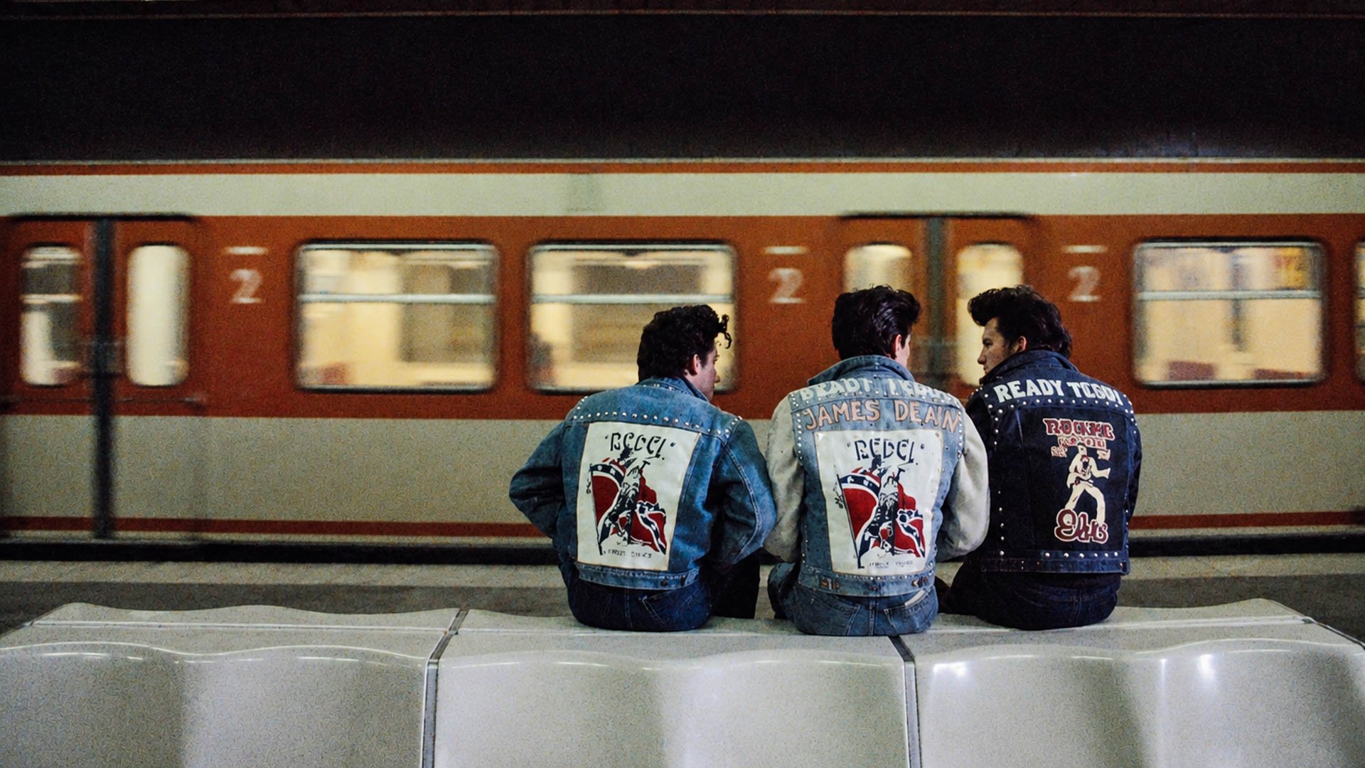 Auf dem Bild sitzen 3 Männer auf einer Bank bei einer U-Bahn Station und es fährt gerade eine U-Bahn ein.