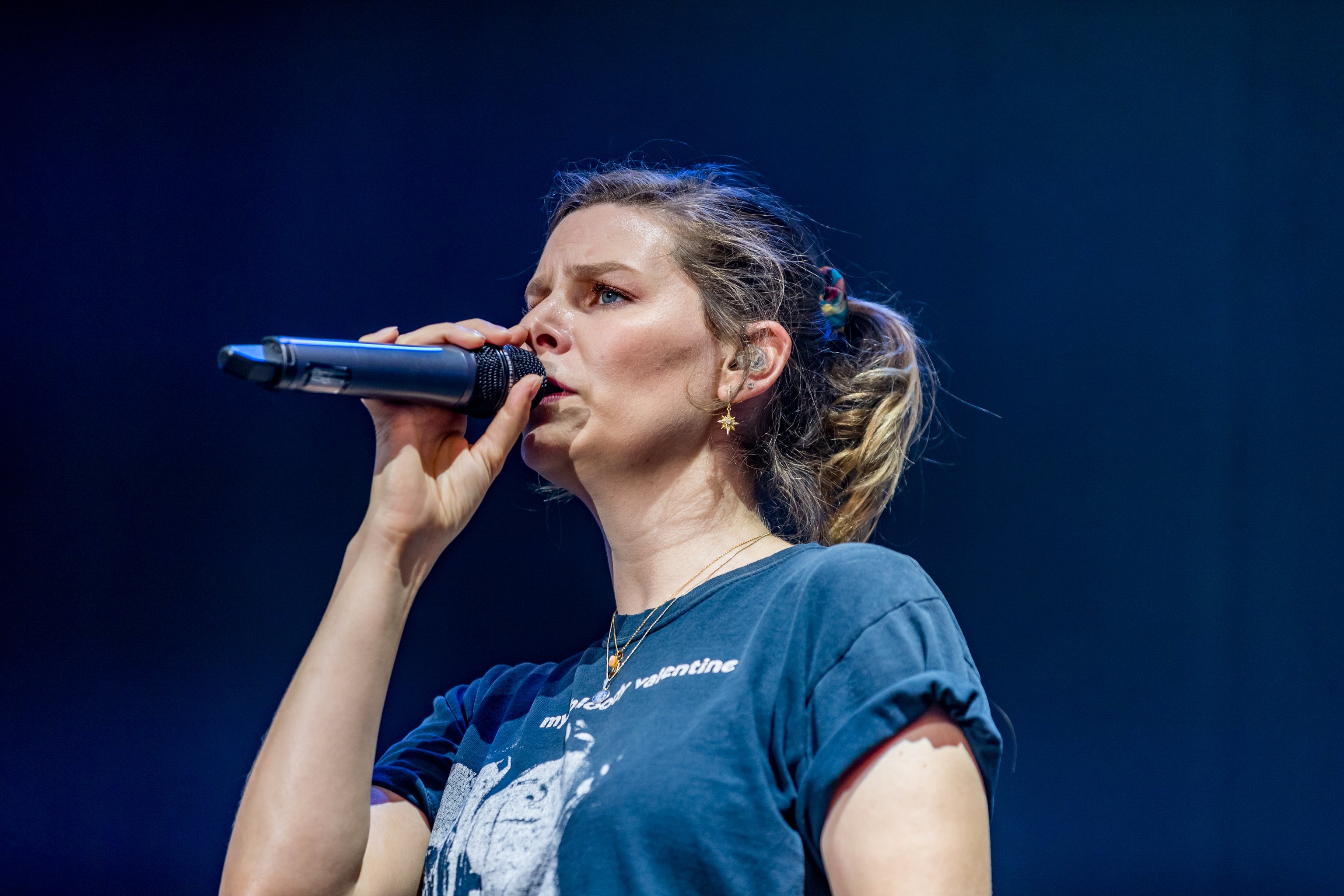 Die Ängerin der Band Juli in einem blauen T-Shirt mit weißem Aufdruck mit einem Mikrofon in der Hand vor blauem Hintergrund
