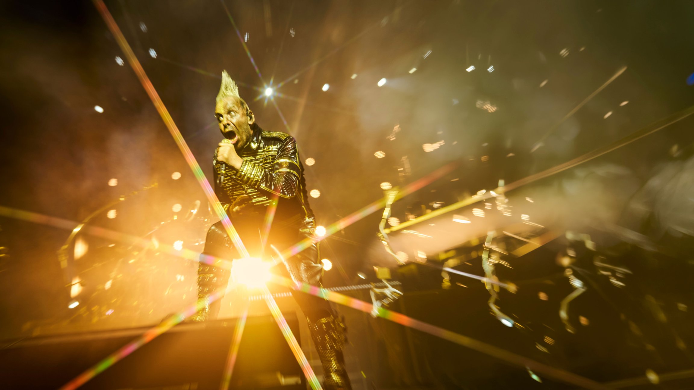 Till Lindemann mit Mikrofon in schwarzem Bühnenoutfit auf einer Konzertbühne. Goldene Lichtstrahlen, Nebel und starke Gegenlichtbeleuchtung prägen die Liveaufnahme.