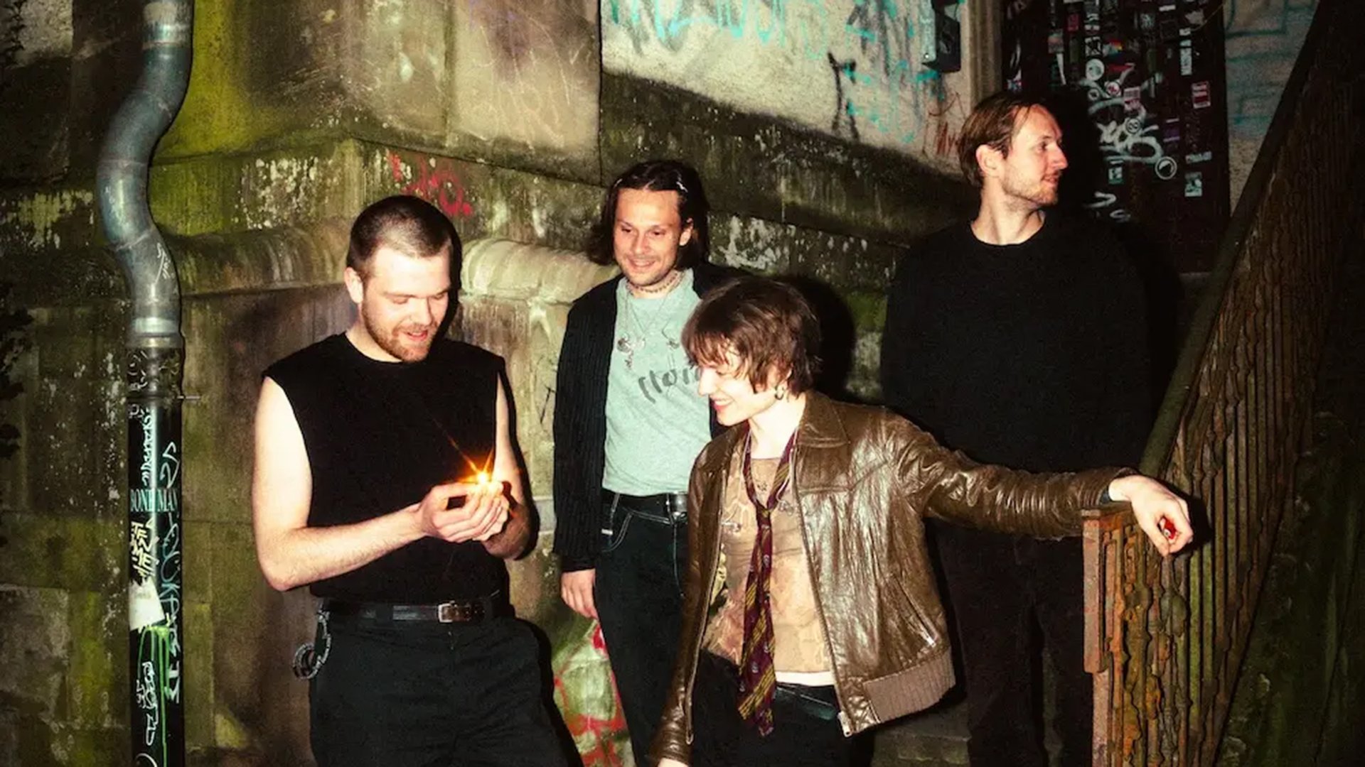 Vier Musiker stehen in urbaner Umgebung mit Graffiti an Wänden, eine Person zündet ein Feuerzeug, alternative Indie‑Band mit Nachtaufnahme und düsterer Atmosphäre