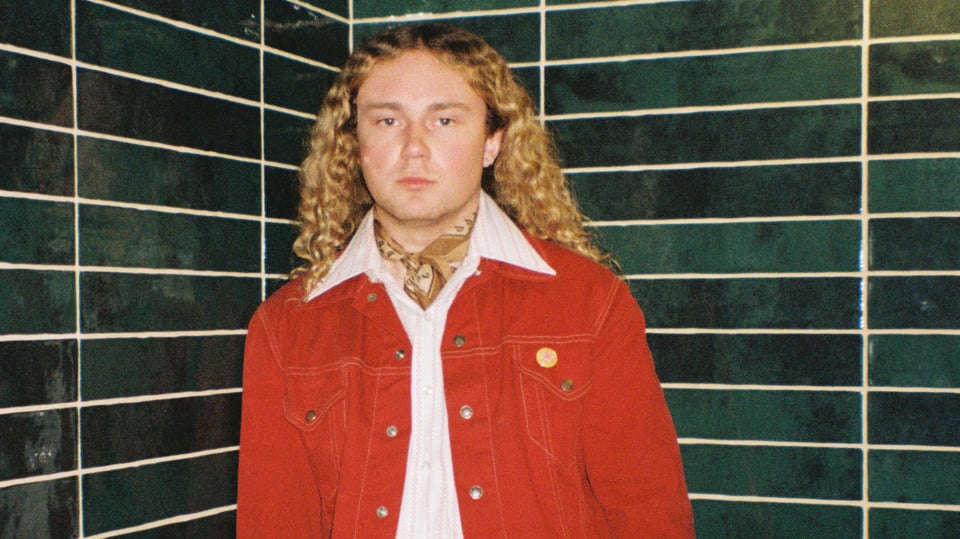 Der Künstler Strawberry Guy in roter Jacke und offenen Haaren vor einer dunkelgrün gefliesten Wand