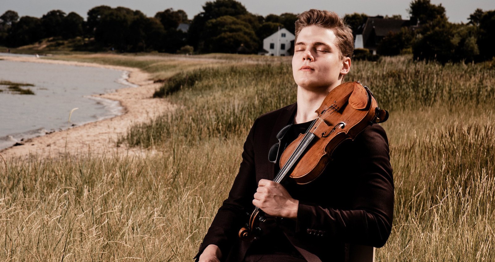 Person mit geschlossenen Augen auf einem Feld sitzend mit Instrument in der Hand