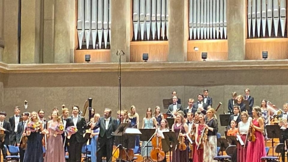 Ein Orchester steht auf einer Bühne, Musikerinnen und Musiker halten ihre Instrumente und Blumen, im Hintergrund eine große Orgel