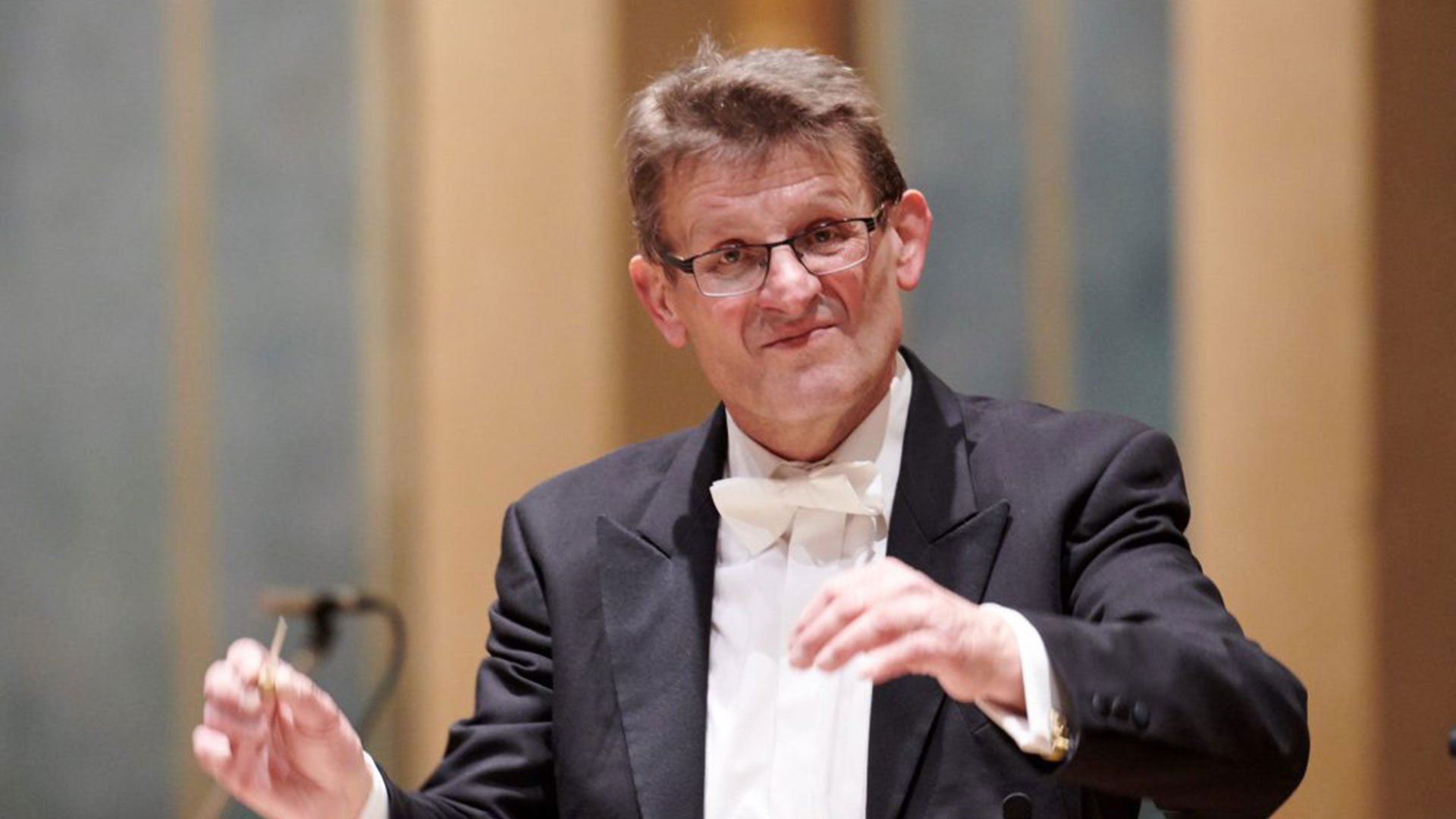 Frühlingskonzert: Dirigent im schwarzen Frack mit weißer Fliege dirigiert auf einer Konzertbühne ein Orchester; im Hintergrund unscharfe Bühnenelemente und ein Instrument.