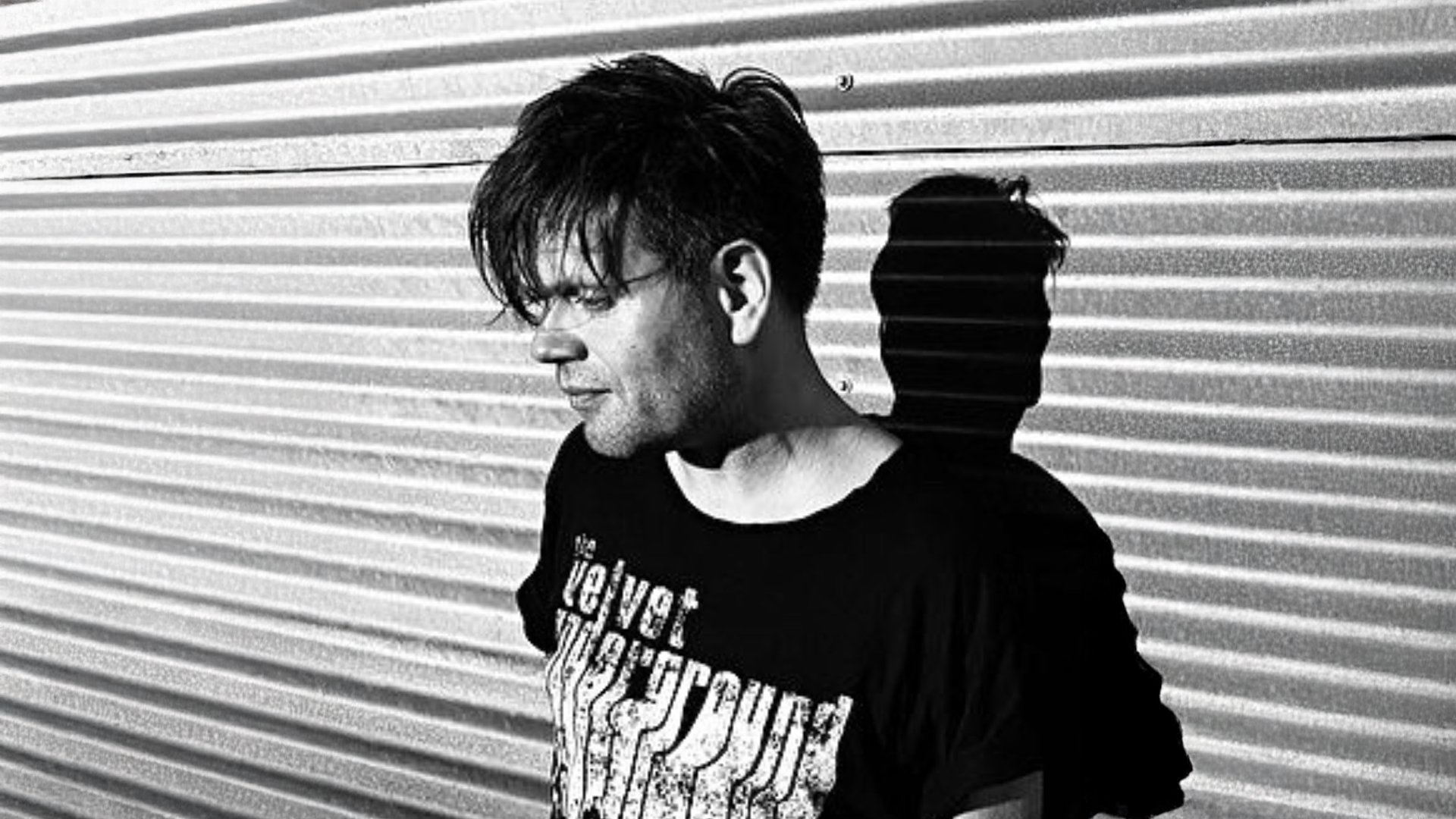 Schwarz‑weißes Porträtmotiv zu Trentemøller: seitliche Ansicht einer Person mit dunklem T‑Shirt vor geriffelter Metallwand, starkes Licht erzeugt einen deutlichen Schlagschatten im Hintergrund; reduzierter, kontrastreicher Look für ein elektronisches Live‑Konzert.