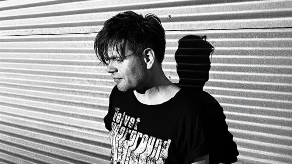 Schwarz‑weißes Porträtmotiv zu Trentemøller: seitliche Ansicht einer Person mit dunklem T‑Shirt vor geriffelter Metallwand, starkes Licht erzeugt einen deutlichen Schlagschatten im Hintergrund; reduzierter, kontrastreicher Look für ein elektronisches Live‑Konzert.