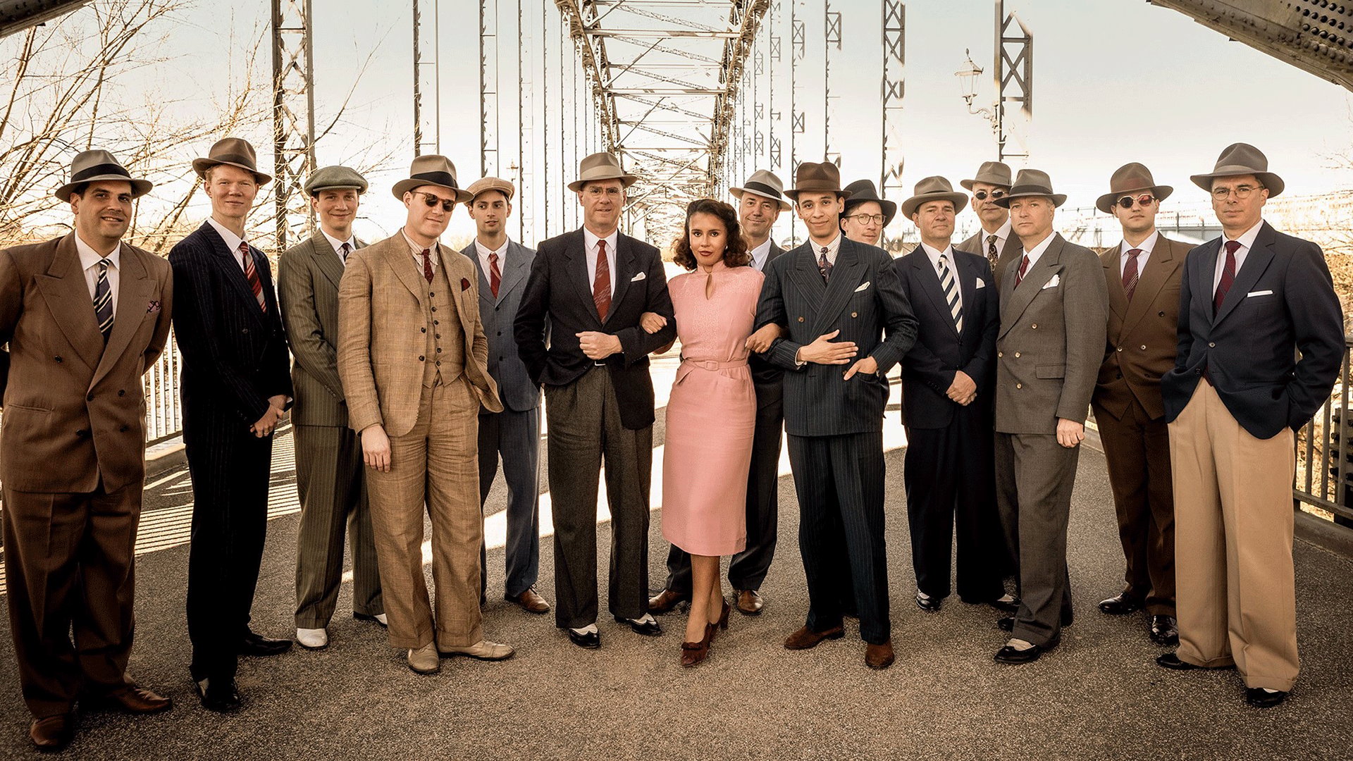 Gruppenfoto eines Swing-Orchesters auf einer Stahlbrücke. Mehrere Personen in Anzügen und Hüten im Stil der 1930er Jahre stehen gemeinsam mit einer Person in rosafarbenem Kleid vor der Brückenkonstruktion und blicken in die Kamera.