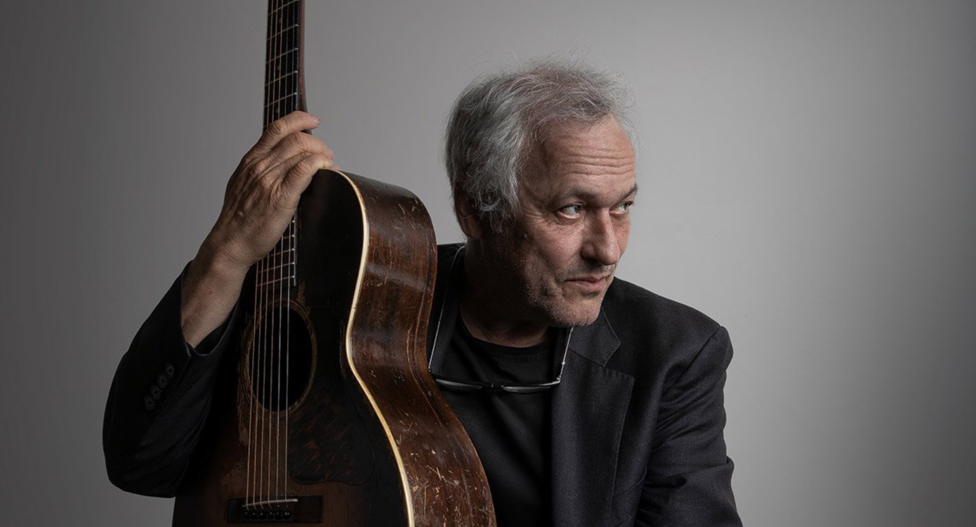 Foto des Künstlers mit Gitarre in den Händen vor grauem Hintergrund