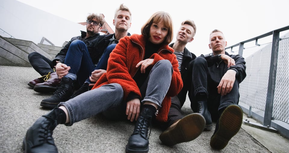 Eine Dame in roter Jacke und vier Herren auf Asphalt sitzend