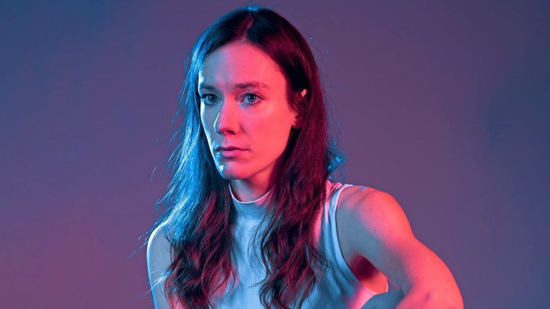 Nahaufnahme einer Person aus Toxische Pommes vor neutralem Hintergrund, seitlich beleuchtet mit blau‑rosafarbenem Licht, mit langem dunklem Haar und hellem ärmellosem Oberteil.