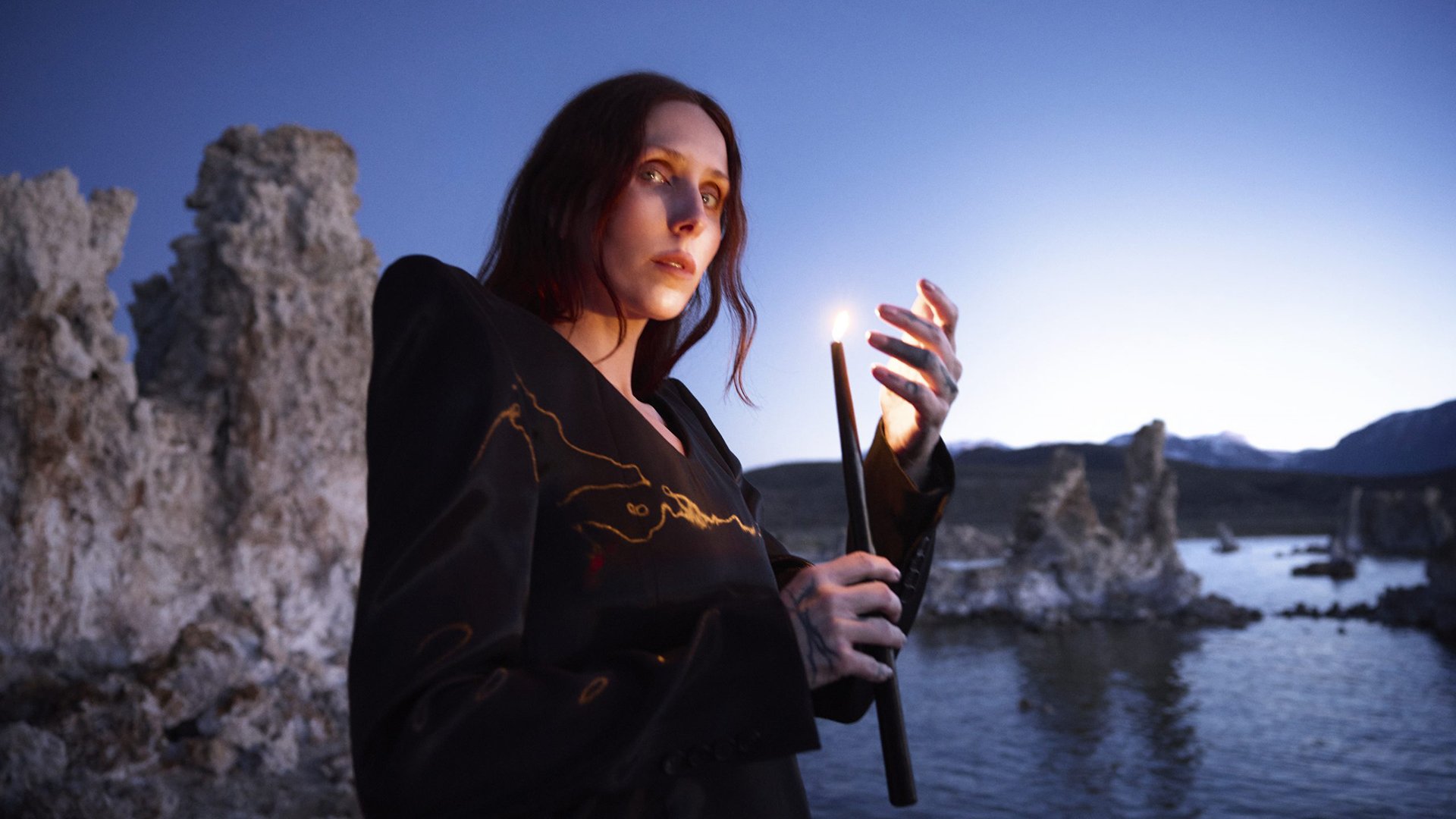 Chelsea Wolfe in schwarzer Jacke steht vor einer felsigen Seenlandschaft bei Dämmerung und hält eine brennende Kerze in der Hand, im Hintergrund Wasser und Steinformationen