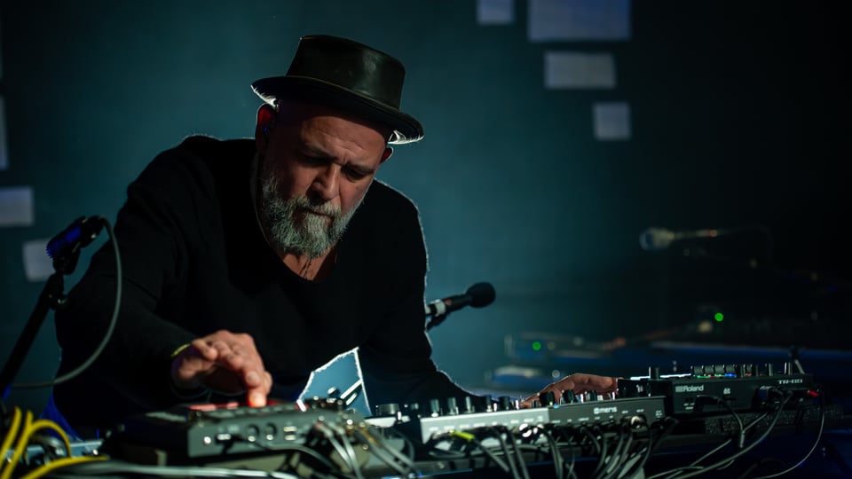 Martin Kälberer mit schwarzem Hut an einem Mischpult und elektronischem Equipment, bedient Regler und Geräte auf einer abgedunkelten Bühne mit blauem Licht und Kabeln im Vordergrund.