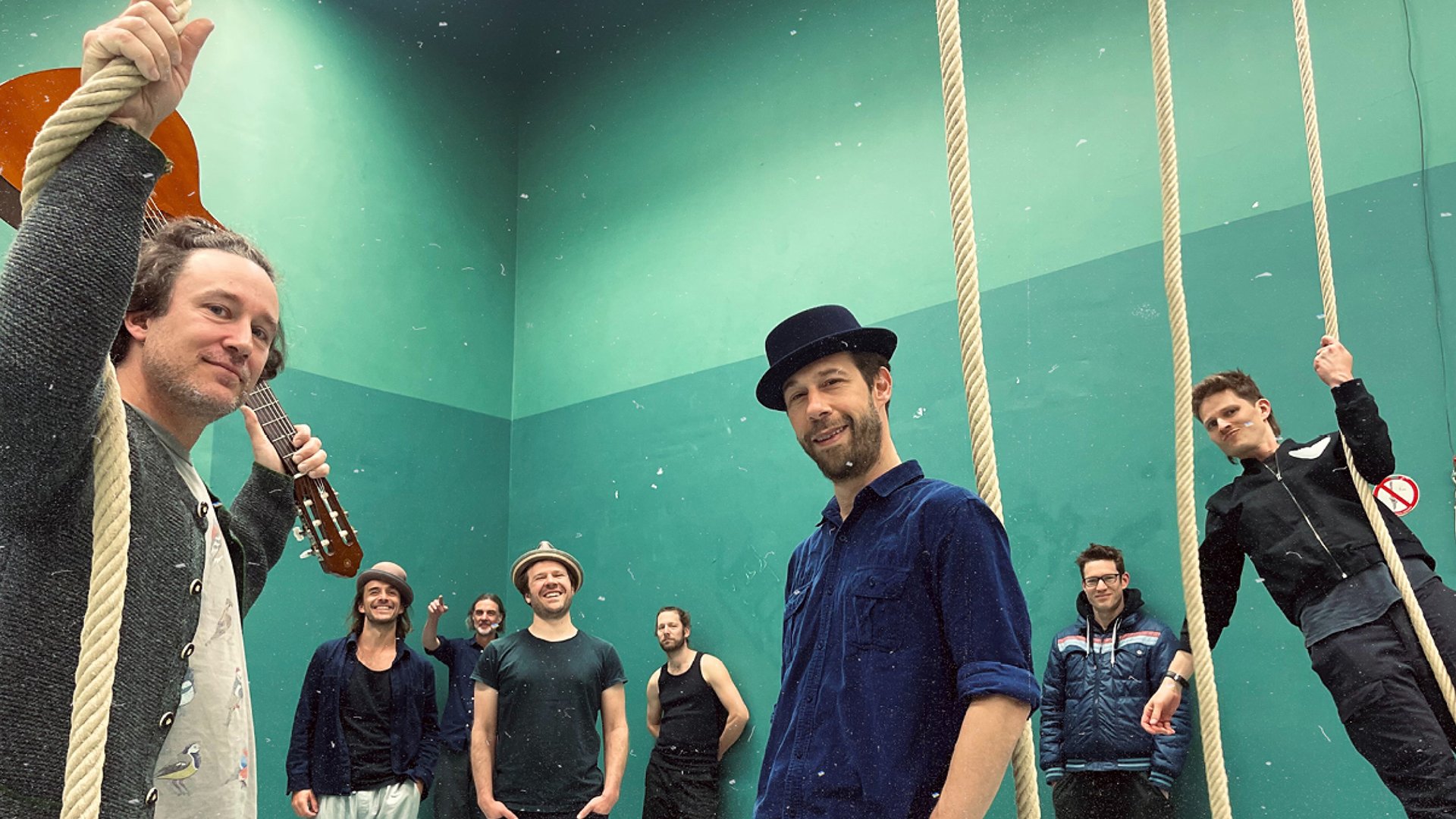 Bandfoto mit acht Personen in einer Halle mit herabhängenden Seilen udn grüner Wand