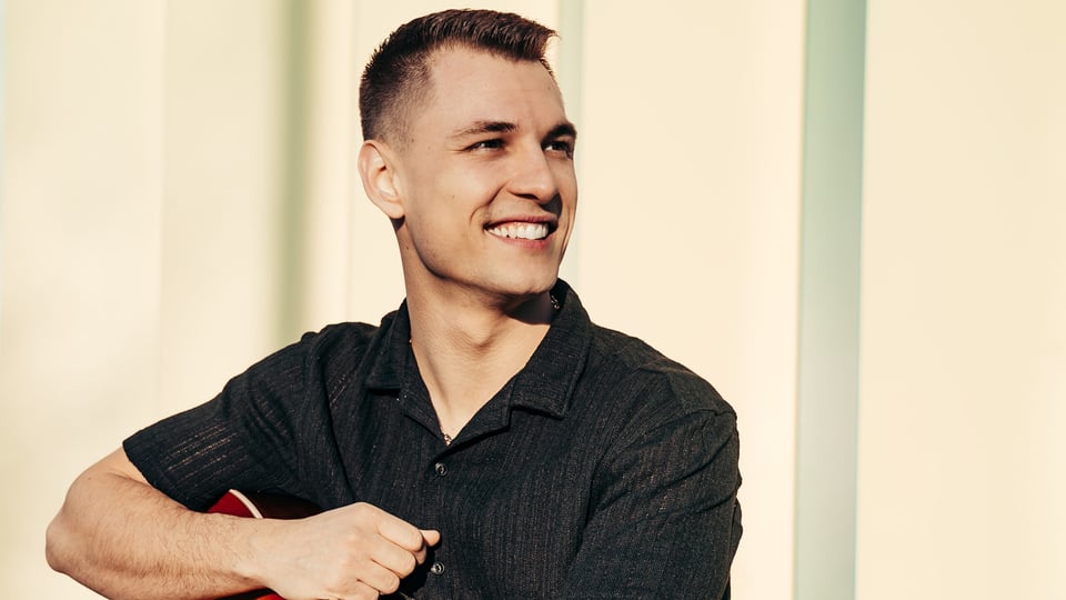 Porträtfoto von Patrick Schneiderwind: Eine Person mit kurz geschnittenen Haaren hält eine Gitarre und blickt seitlich aus dem Bild. Die Person trägt ein dunkles Hemd; der Hintergrund ist hell und reduziert gestaltet. Die Aufnahme ist weich ausgeleuchtet und dient als Künstler‑ und Pressefoto für musikalische Veranstaltungen.