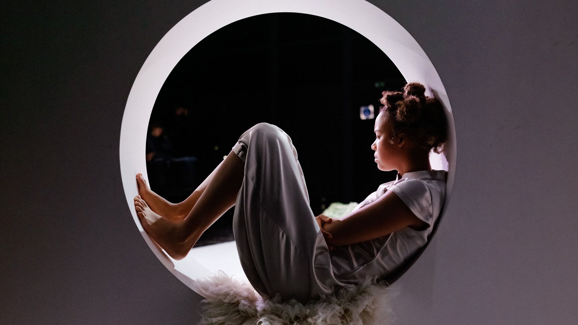 Kind sitzt mit angezogenen Beinen in einer runden Öffnung auf einer Bühne, vor dunklem Hintergrund und weichem Licht.