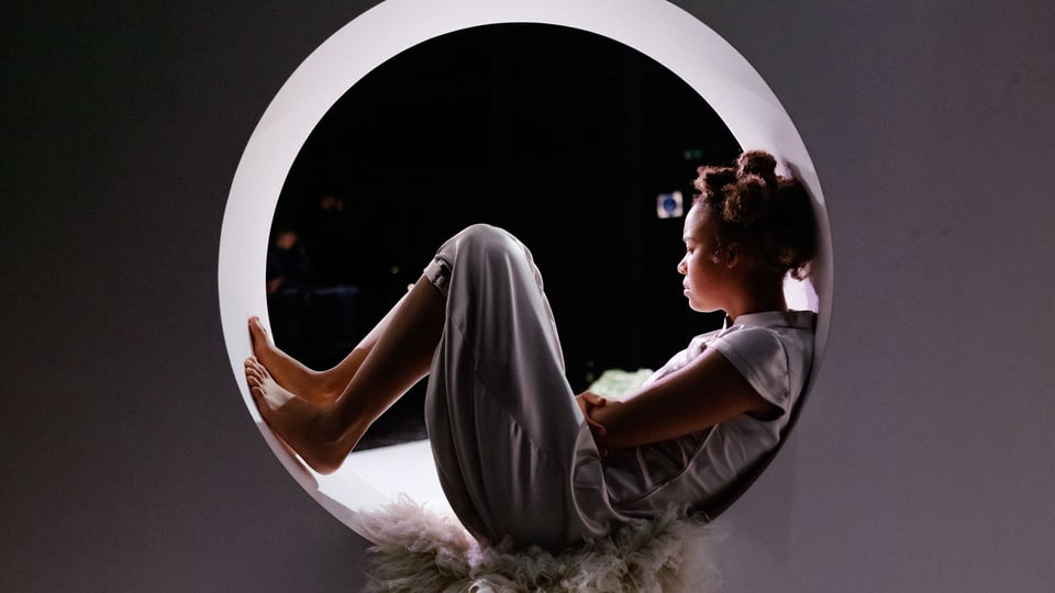 Kind sitzt mit angezogenen Beinen in einer runden Öffnung auf einer Bühne, vor dunklem Hintergrund und weichem Licht.