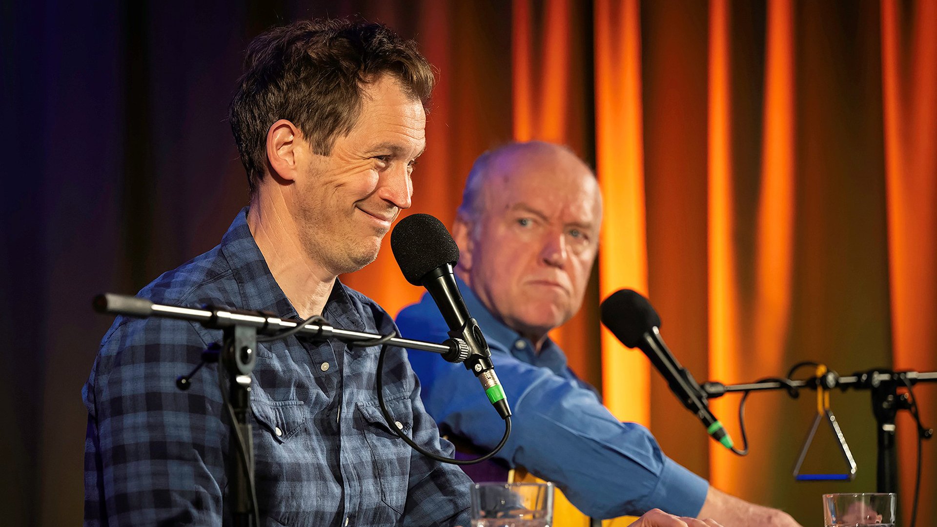 Bühnenfoto mit zwei Personen an Mikrofonen auf einer Bühne vor orange beleuchtetem Vorhang, eine Person im blau karierten Hemd im Vordergrund, die andere in blauem Hemd dahinter, vor Mikrofonständern und einem Glas auf dem Tisch.